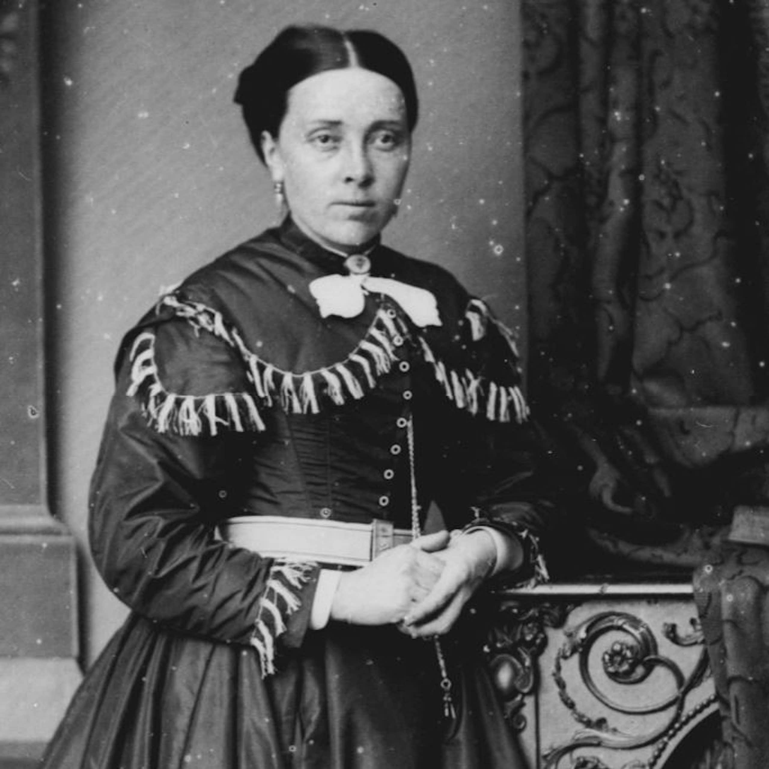 A black and white photo of a Victorian looking woman with her hands clasped. She wears a heavy dress with tassles across the chest area. Her hair is pinned back tightly. 