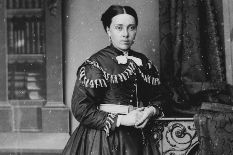 A black and white photo of a Victorian looking woman with her hands clasped. She wears a heavy dress with tassles across the chest area. Her hair is pinned back tightly.