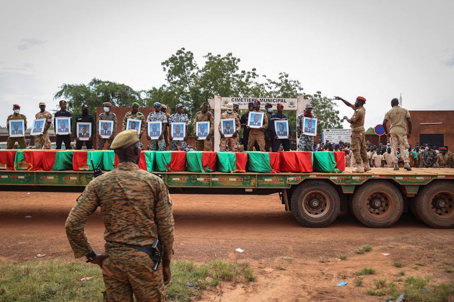 Men wearing military uniforms kneel on coffins draped in red and green that are on the back of a truck