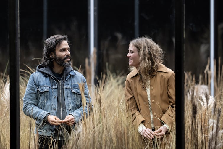 Sofiane and Isolde stand next to each other in a glass box full of wheat plants.