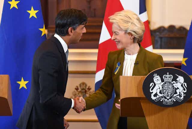 Rishi Sunak and Ursula von der Leyen look at each other and shake hands, smiling, while standing behind a podium and in front of an EU flag and UK flag