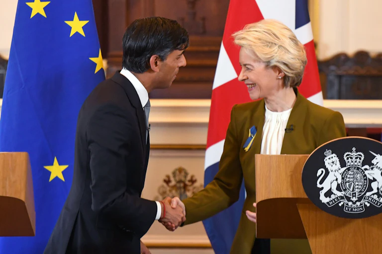 Rishi Sunak and Ursula von der Leyen look at each other and shake hands, smiling, while standing behind a podium and in front of an EU flag and UK flag