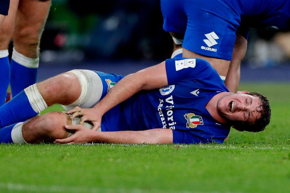 A man wearing a blue Italian rugby kit lies on the floor clutching his leg. He appears to be in pain and has his mouth open. 