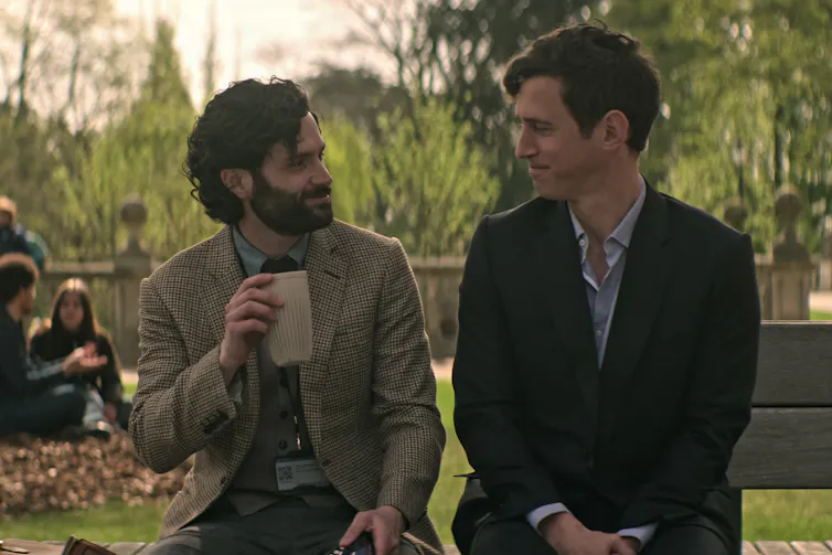 Penn Badgley as Joe Goldberg, Stephen Hagan as Malcolm sit on a bench outside the university.