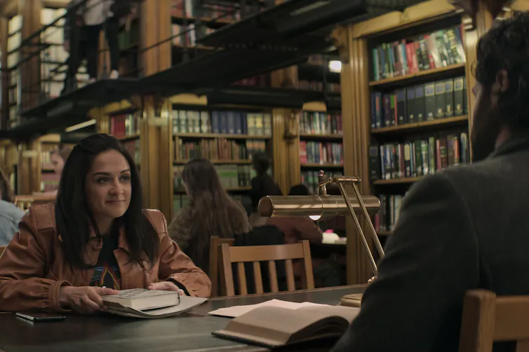 A student sits in a university library smiling at a man off camera.