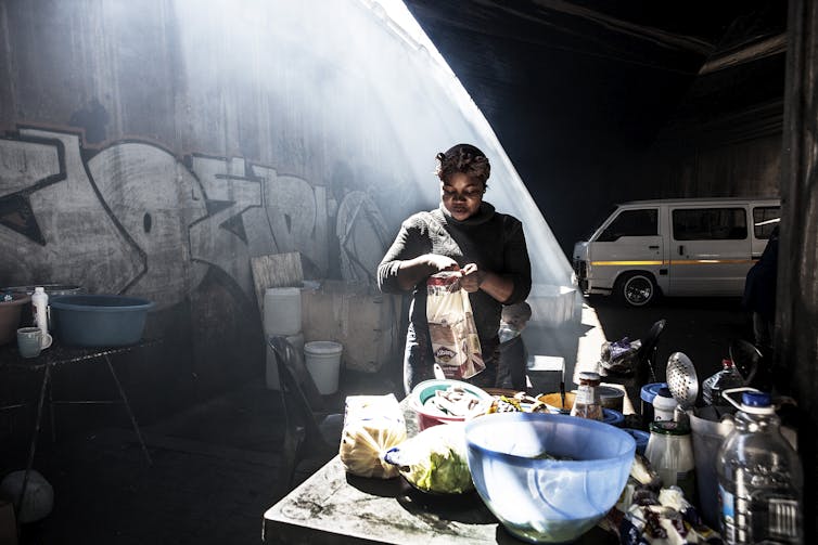 A woman stands in a dark space under a bridge in a beam of bright light, taking bread from a bag behind a table used for food preparation.