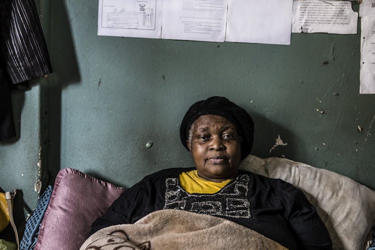 An older woman rests in bed, looking directly at the camera without smiling, papers stuck to the wall above her.