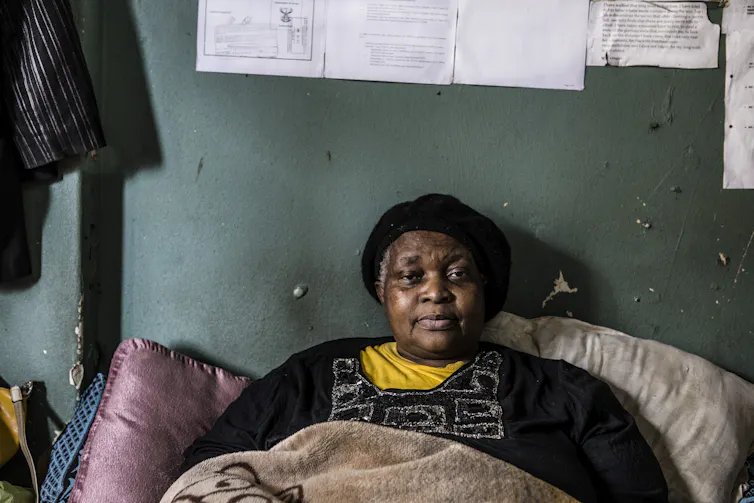 An older woman rests in bed, looking directly at the camera without smiling, papers stuck to the wall above her.