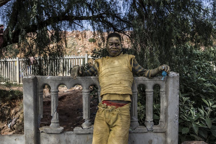 A man with a spry expression leans against a broken balustrade, his tattered clothes covered in dust.