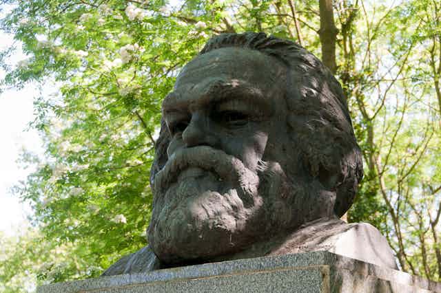 Karl Marx's bust in Highgate Cemetery, London with trees in bloom behind it.
