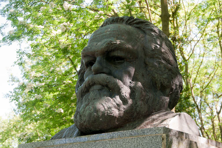 Karl Marx's bust in Highgate Cemetery, London with trees in bloom behind it.