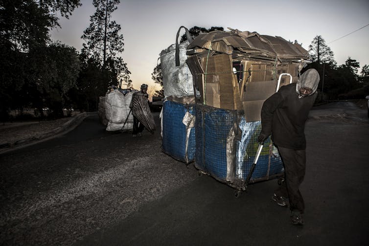 In dim light, men pull trolleys with shiny containers loaded with cardboard.