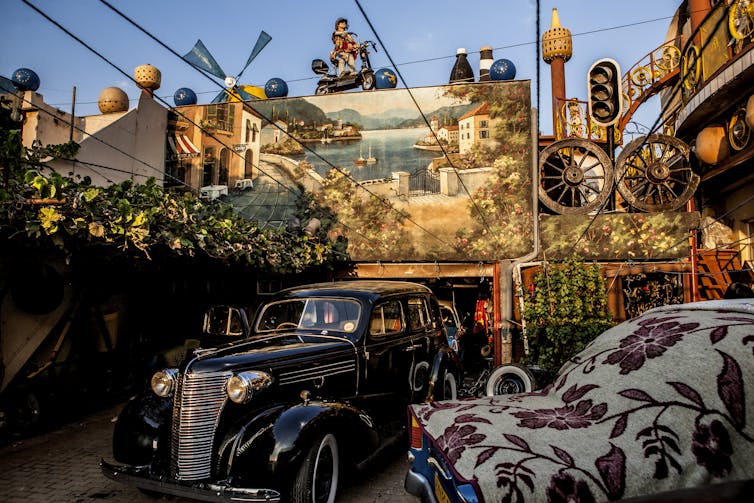 A visually rich exterior of a house with vintage cars, a mural of a town near water, a windmill, a statue of a tower, concrete wagon wheels and creepers.