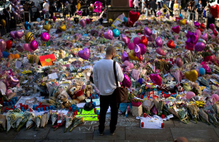 A man standing in front of flowers and toys laid in memory of the victims of the Manchester Arena bombing in 2017.