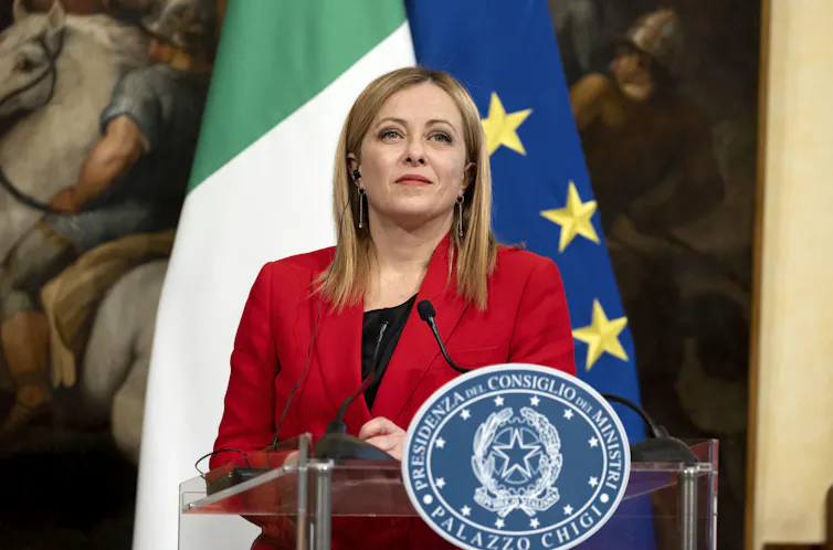 Giorgia Meloni at a podium in front of Italian and EU flags.