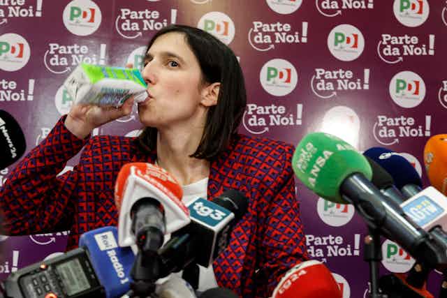 Elly Schlein swigs from a drink carton in front of microphones at a press conference. 