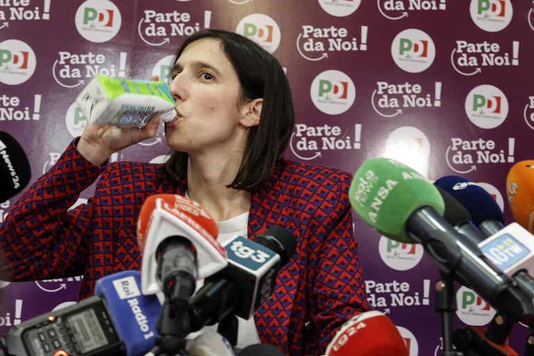 Elly Schlein swigs from a drink carton in front of microphones at a press conference.