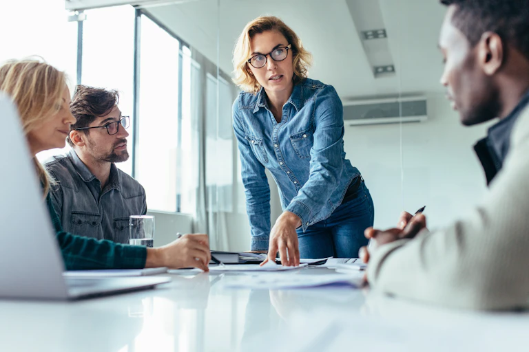 A young woman stands at the head of a conference table and explains something to colleagues seated around