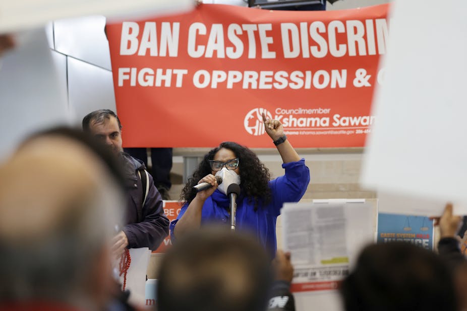 A woman dressed in a blue shirt speaking to an audience sitting in front of her with a large poster saying 'Ban caste discrimination' behind her.