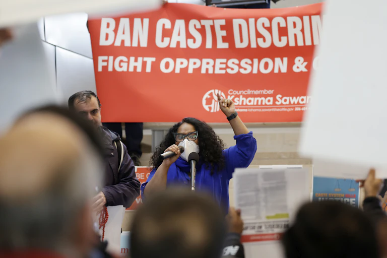 A woman dressed in a blue shirt speaking to an audience sitting in front of her with a large poster saying 'Ban caste discrimination' behind her.