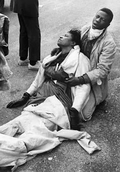 A man crouching on the pavement cradles an injured woman.