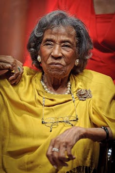 A sitting woman with gray hair in a gold-colored dress and jewelry.