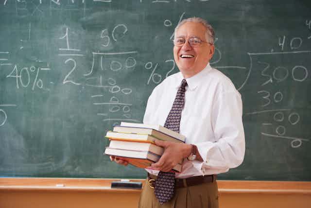 An older male teacher holds a stack of books while standing in front of a green chalkboard. He is smiling.