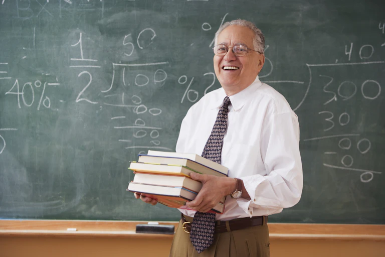 An older male teacher holds a stack of books while standing in front of a green chalkboard. He is smiling.