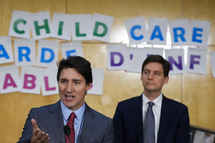 A man speaking while another stands behind him. The words child care appear on the wall behind them.