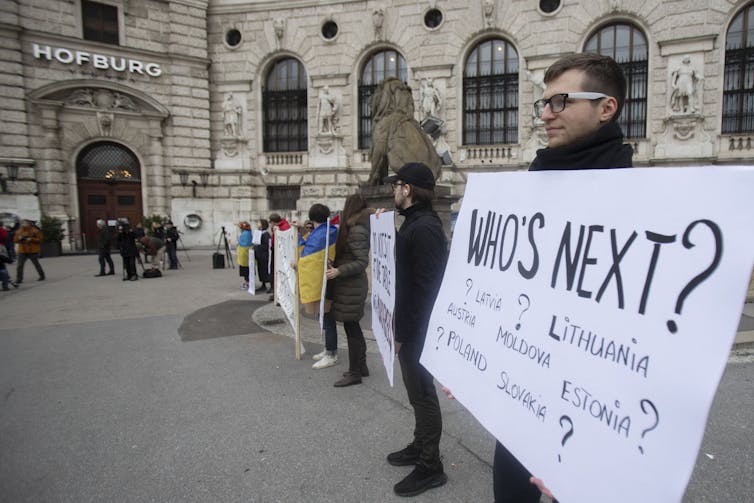 Un hombre sostiene un cartel que dice '¿quién sigue?  letonia?  ¿Lituania?  Moldavia?  Está parado en lo que parece una antigua calle europea con un gran edificio detrás de él.