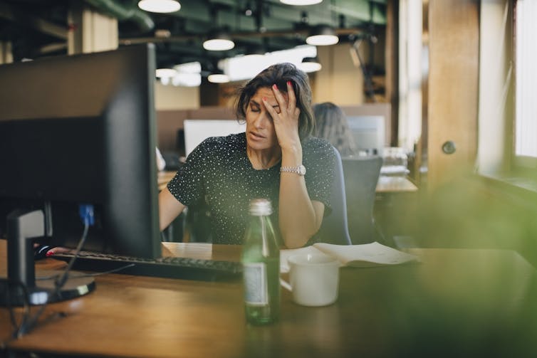 a woman seated at a desk in front of a computer with her eyes closed and her left hand on her forehead