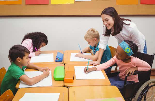 A teachers assists a group of children at a desk. One of them is sitting in a wheelchair.