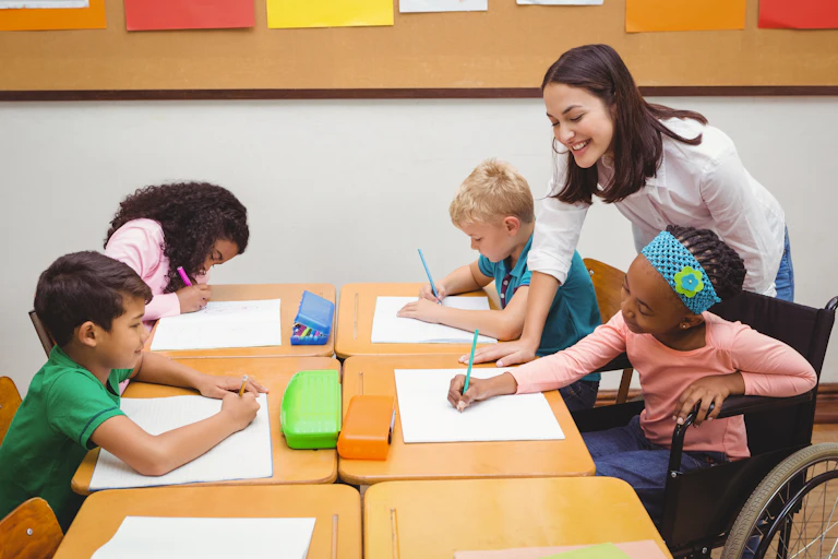 A teachers assists a group of children at a desk. One of them is sitting in a wheelchair.