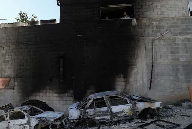 Two burnt cars stand next to a wall blackened by fire while some people look at the scene. 