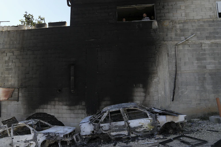 Two burnt cars stand next to a wall blackened by fire while some people look at the scene.