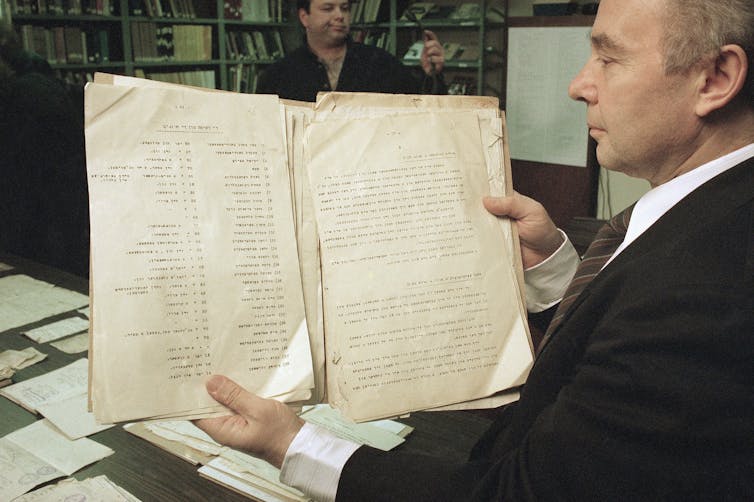 A man wearing a black jacket holding loose sheets of papers with lists of names on them.