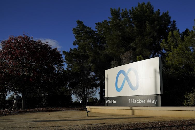 billboard showing an infinity loop in blue on a white background sits next to a road as a person walks past with trees in distance