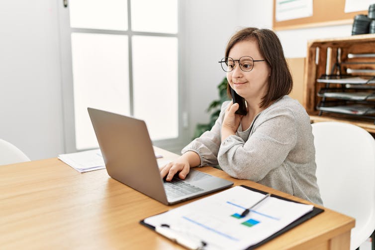A woman with down syndrome sits at a table in a bright office using laptop.