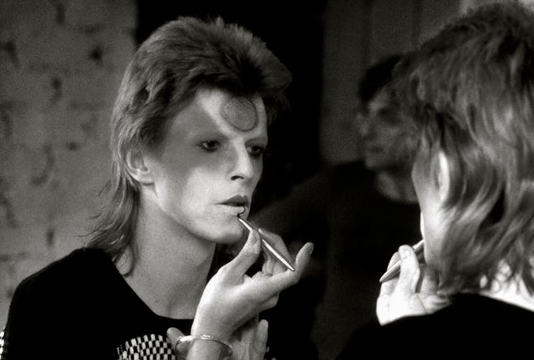 David Bowie puts on makeup in the mirror.