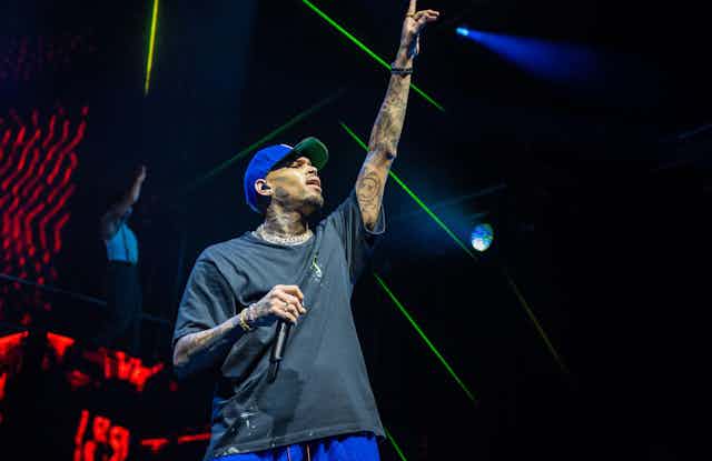 Chris Brown on stage wearing a grey t-shirt and blue cap. He holds a microphone and one arm is aloft.