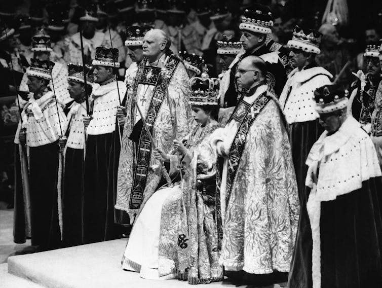 A black and while photo of a woman wearing a crown sitting on a throne with men standing around her.