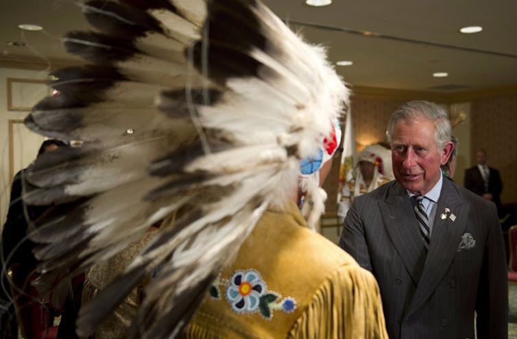 Charles shakes hands with an Indigenous man wearing a traditional feather headdress.
