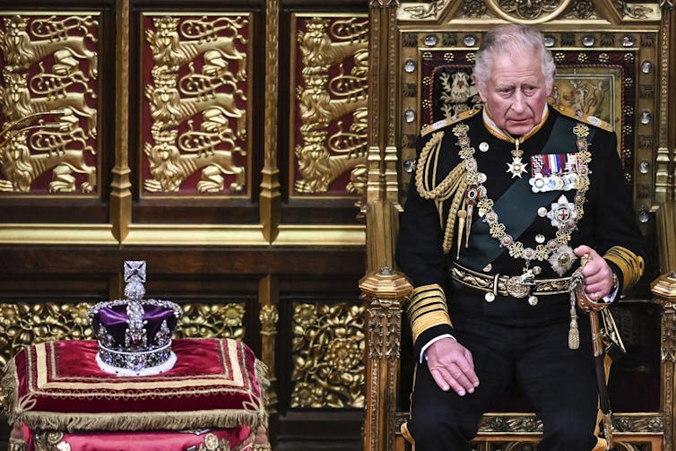 Charles in a military uniform sitting on a gold throne next to a bejeweled crown of a red pillow.