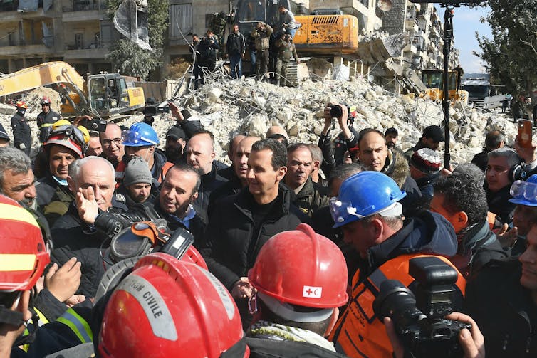 A man dressed in dark clothes is surrounded by other men wearing helmets as they walk near a demolished building.