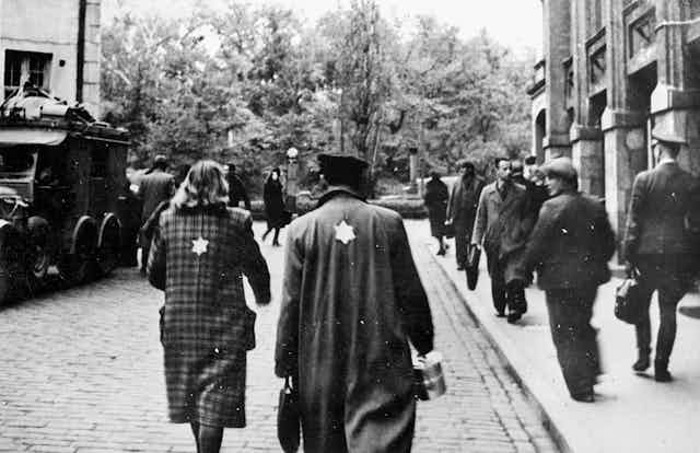 A man and woman walk down a cobblestone street, wearing coats with yellow Stars of David on the back.