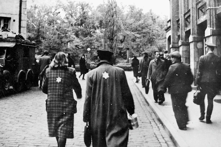 A man and woman walk down a cobblestone street, wearing coats with yellow Stars of David on the back.