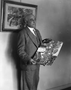 A black and white photograph of a bald man in a suit holding a painting.