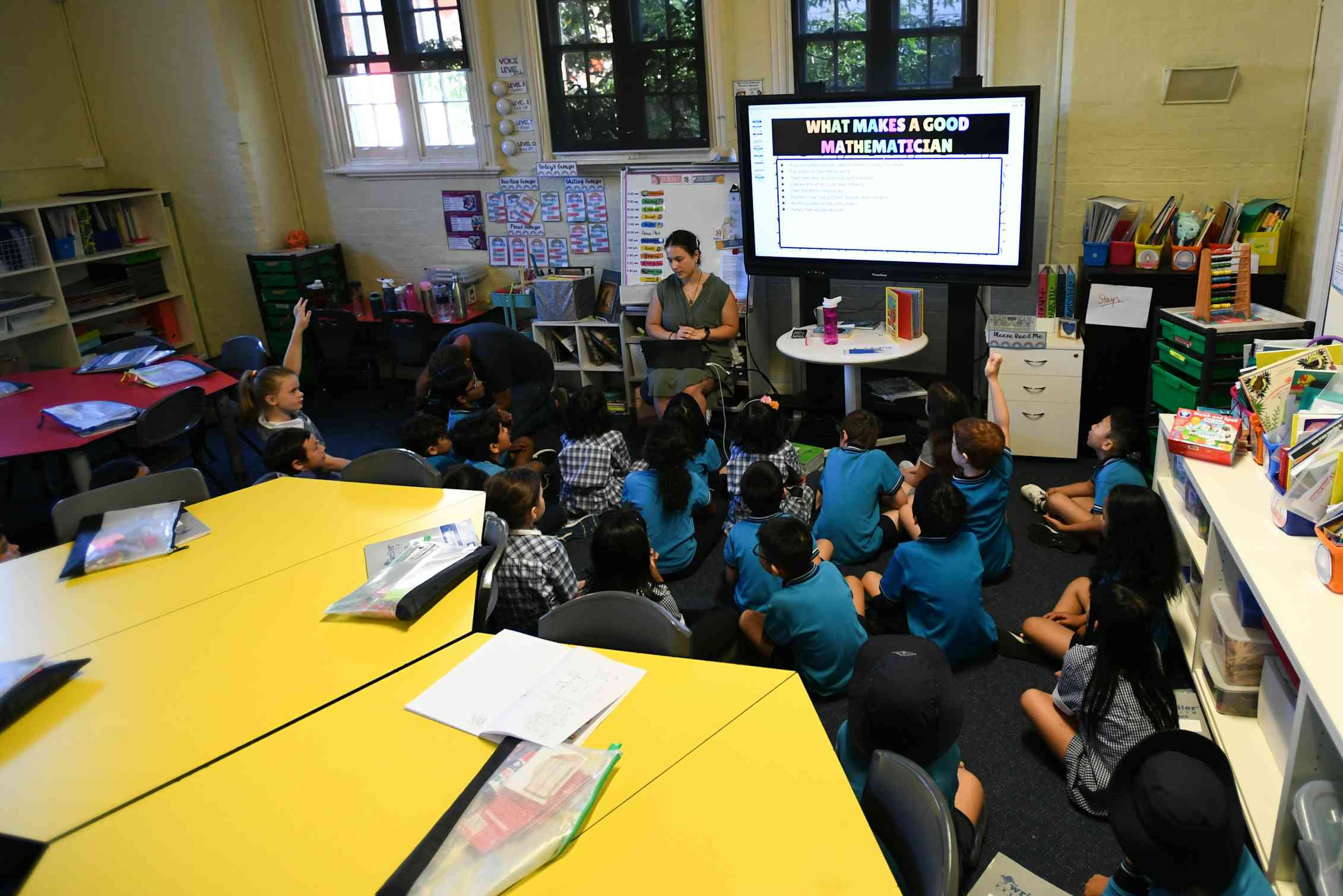 Primary students sit on the classroom floor, listening to their teacher.