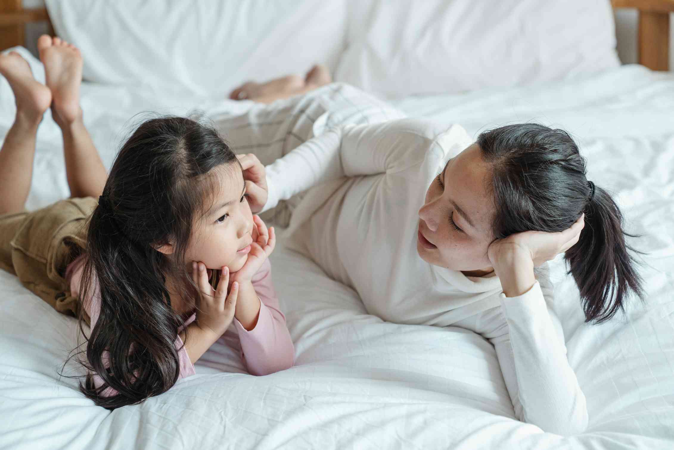 A mother and young daughter, lying on a bed and talking.