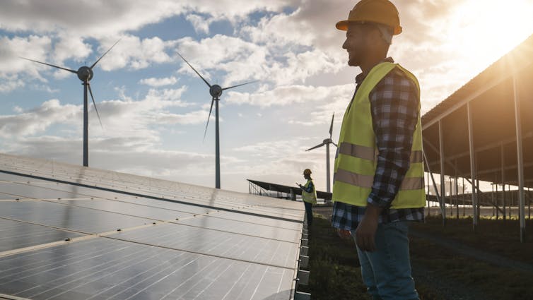 Man wearing a hi-vis vest and hard hat looking at solar panels with wind turbines in the background.